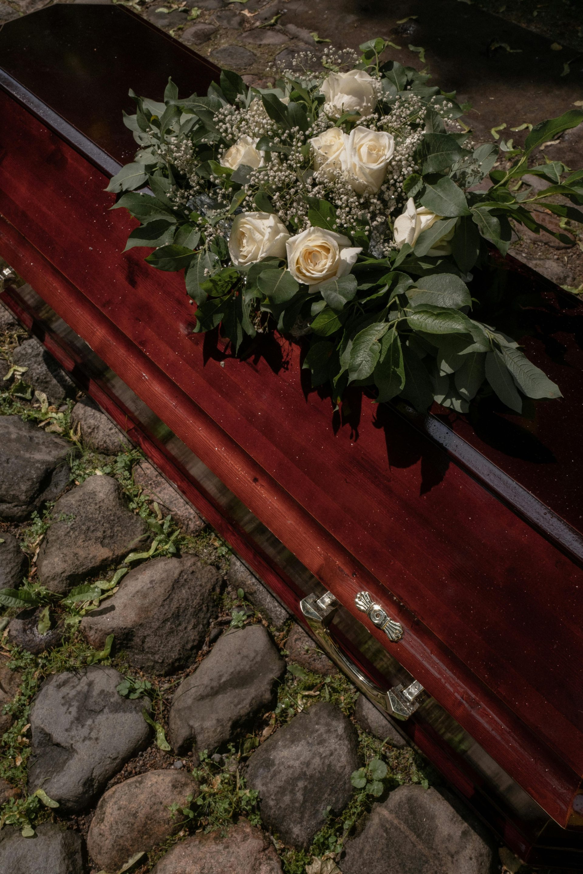 Ein Mahagonisarg, geschmückt mit einem üppigen Kranz aus weißen Rosen, steht auf einem friedlichen Friedhof.